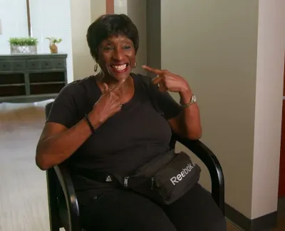 Smiling woman sitting in a dental office pointing at her teeth, wearing a black shirt and Reebok fanny pack.
