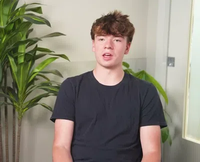 Teenage boy in a black t-shirt sitting indoors next to green potted plants and a sign reading You Make Me Smile.