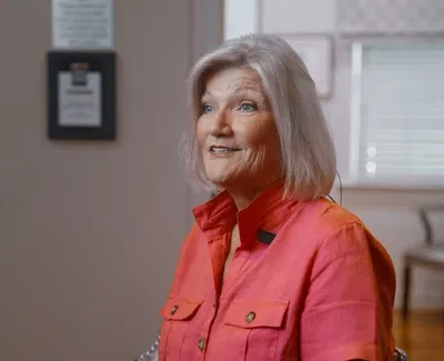 Smiling elderly woman with short white hair wearing a coral button-up shirt indoors with soft natural light.