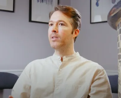 Man in white shirt speaking in a modern living room with orchids and framed artwork on the wall.
