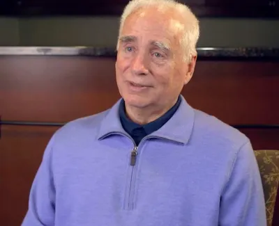 Elderly man in a light purple zip-up sweater sitting indoors with a neutral expression against a wooden background.