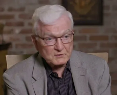 Elderly man with white hair and glasses speaking against a brick wall background in a formal setting