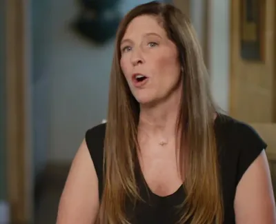 Woman with long brown hair speaking indoors, wearing a black sleeveless top, sitting on a chair.
