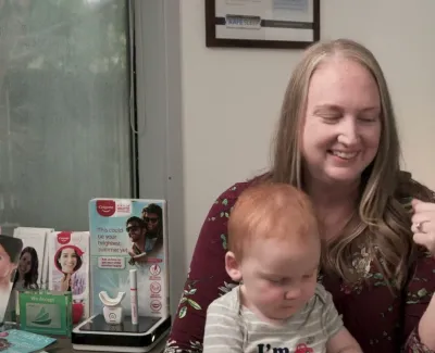 Smiling woman holding a toddler in a dental office with informational flyers and service list in background