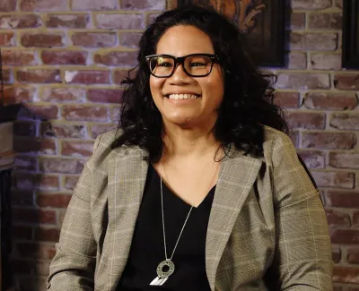 Smiling woman with glasses and curly hair sits in front of brick wall and wooden cabinet indoors.