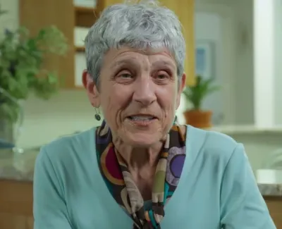 Elderly woman with gray hair and patterned scarf smiling indoors in a cozy, softly lit room.