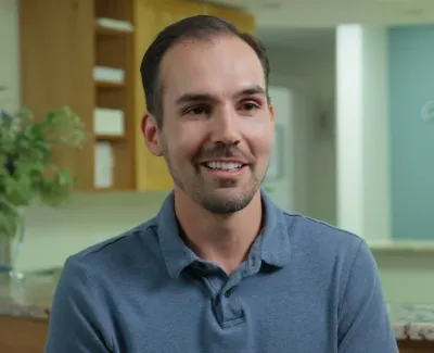 Man in a blue polo shirt smiling in a modern office with a plant and blurred background decor.