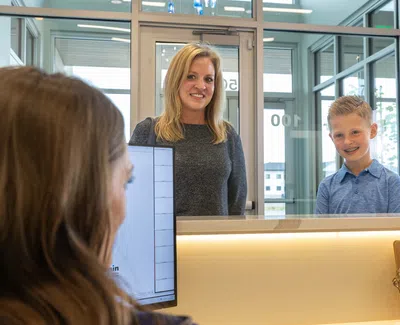 Woman and young boy smiling at reception desk inside modern building with glass doors and windows.