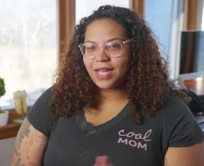 Woman with curly hair and glasses wearing a cool mom t-shirt in a cozy office setting with a window behind.