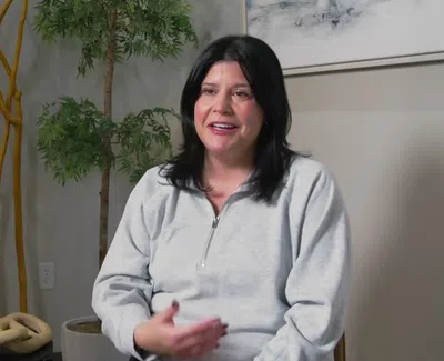 Smiling woman in gray sweatshirt sitting indoors with green plants and decorative wooden branches in the background