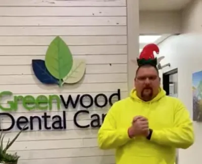 Man in bright yellow hoodie and elf hat standing inside Greenwood Dental Care office with logo on white wall.