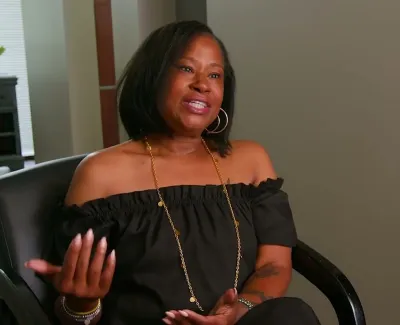 Smiling woman in black off-shoulder top sitting and gesturing during an indoor interview or conversation.