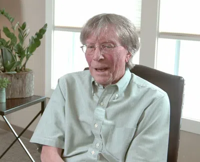 Elderly man with glasses sitting in a chair inside a well-lit living room with plants and a lamp.