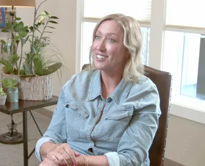 Smiling woman in denim shirt sitting in a cozy living room with plants and natural light from windows
