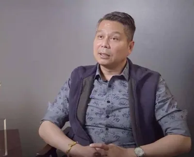 Man in patterned shirt and vest sitting in chair beside table with plant and award plaque in office.