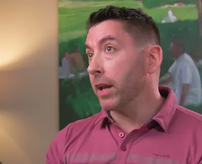 Man in a pink shirt speaking indoors with a modern lamp and green painting in the background