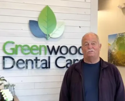 Older man standing inside Greenwood Dental Care with green leaf logo on white wooden wall behind him