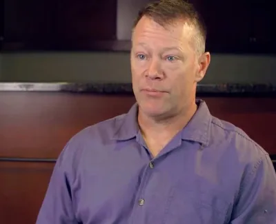 Man in a purple shirt speaking indoors with a neutral brown wall and dark cabinets in the background