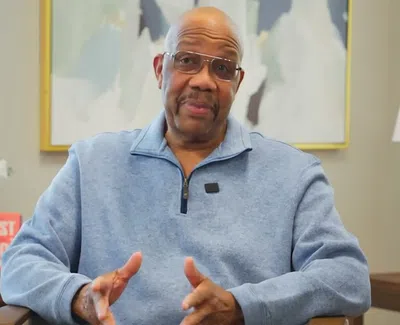 Man in glasses and blue sweater speaking in a well-lit room with artwork and a lamp in the background