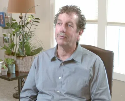 Man with curly hair and mustache sitting in a chair in a living room with plants and a lamp behind him.