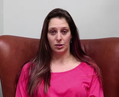 Woman with long brown hair wearing a pink shirt sitting on a brown leather chair against a plain wall with a plant nearby.