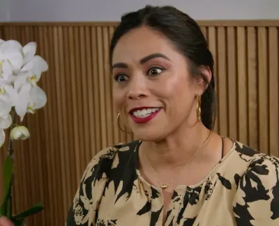 Smiling woman in black and beige blouse with gold hoop earrings sitting near a white orchid and wooden wall paneling.