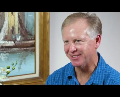 Smiling man in blue checkered shirt sitting beside framed sailing ship painting and white orchid flowers.