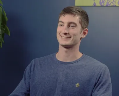 Young man smiling sitting indoors next to a tall leafy plant against a blue wall background