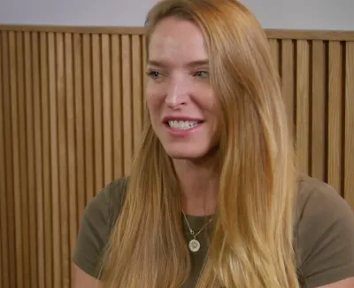 Smiling woman with long blonde hair wearing a necklace and green shirt sitting by a wooden slat wall with white orchids.