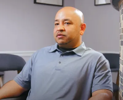Man in gray polo shirt sitting in a living room with gray chairs and framed art on the wall.
