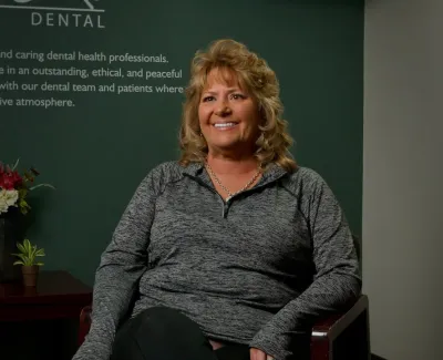 Smiling woman sitting in dental office with inspirational dental care message on wall behind her