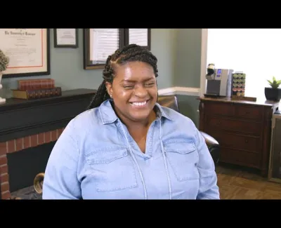 Smiling woman with braided hair in a denim shirt sitting in a cozy office with framed certificates and wooden furniture.