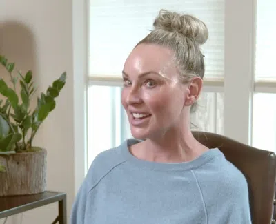 Smiling woman with blonde bun sitting in chair near window and indoor plants on side table.