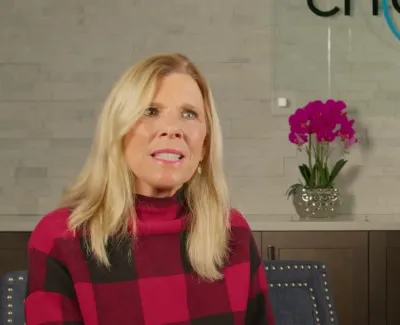 Blonde woman in red checkered sweater sitting in modern dental office with orchid and logo behind her