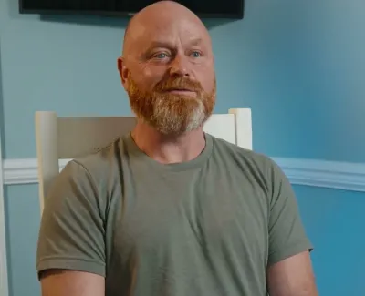 Bearded man in green t-shirt sitting on white chair in blue room with plant in corner