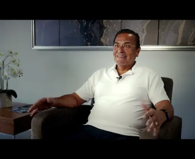 Man in white polo shirt sitting in an armchair with a table and orchid plant in the background indoors.