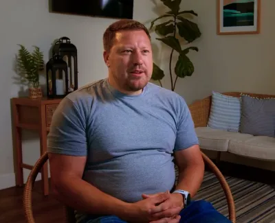 Man in a blue t-shirt sitting on a wicker chair in a cozy living room with soft lighting and plants.
