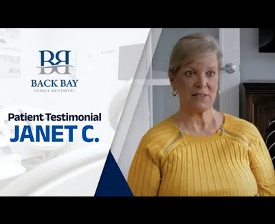 Patient Janet C. giving testimonial for Back Bay Family Dentistry, wearing a yellow top in a home setting.