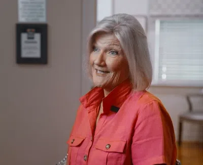Smiling elderly woman with white hair wearing a bright pink shirt sitting indoors near a window with blinds.