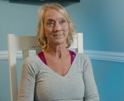 Smiling middle-aged woman with blonde hair sitting in a chair against a blue wall with a plant nearby.