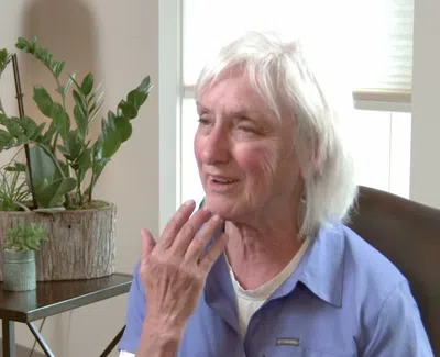 Elderly woman with white hair smiling and touching her face while sitting indoors near plants and a lamp.