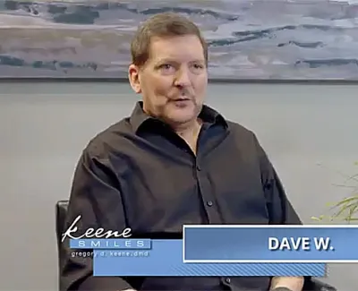 Middle-aged man in a black shirt giving a testimonial in a dental office setting with a plant and artwork behind.