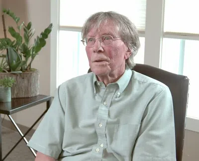 Elderly man with glasses sitting in chair near table with potted plants in bright room with large window.