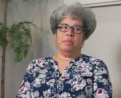 Older woman with glasses and curly gray hair wearing floral blouse sitting indoors with plants behind her