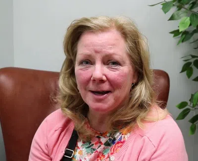 Smiling middle-aged woman with blonde hair wearing a floral top and pink cardigan sitting on a brown chair indoors.