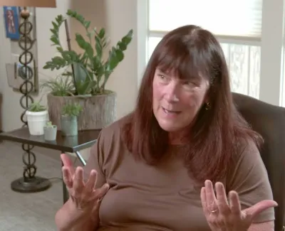 Woman with long brown hair talking animatedly while seated in a living room with plants and natural light.