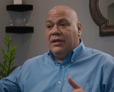 Bald man in blue shirt gesturing while speaking in a modern indoor setting with plants and shelves.