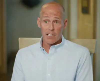Bald middle-aged man in a light blue shirt speaking indoors with a blurred background.