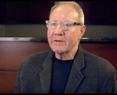 Elderly man wearing glasses and a dark blazer speaking in an indoor setting with a blurred background.