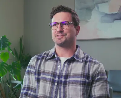 Man wearing glasses and plaid shirt sitting indoors near a window with plants and abstract art on the wall.